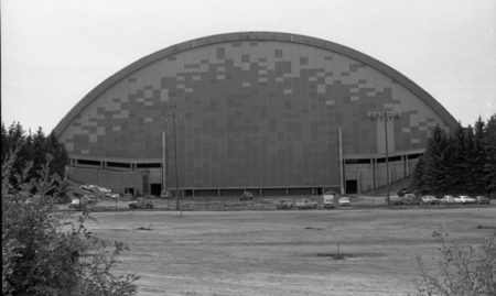 Black and white photograph of an arched building, its end wall decorated with different shades. In the foreground are several vehicles.