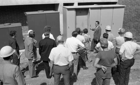 Black and white photograph of a group of people standing next to metal boxes on the side of a building.