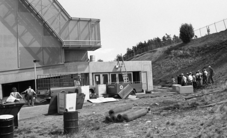 Black and white photograph of a group of people standing on a hillside next to a concrete building.
