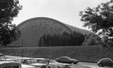 Black and white photograph of a building with an arched roof and speckled decor on one end wall. Cars are parked along a street in the foreground.