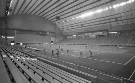 Black and white photographs of the interior of an enclosed stadium. Football players stand around the football field. The stands are empty.