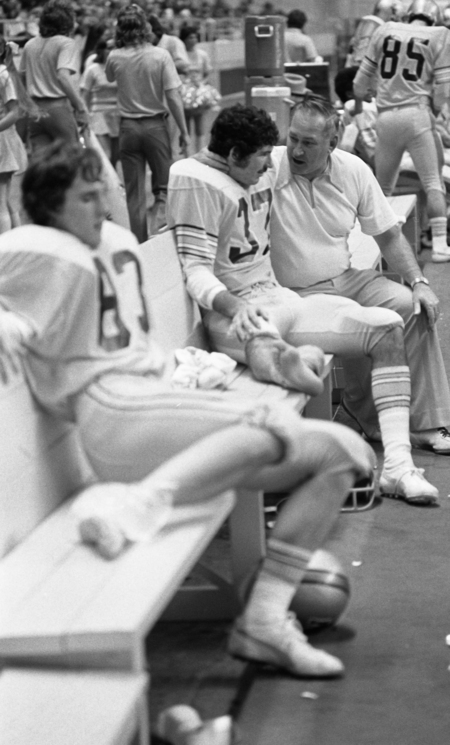 Black and white photograph of a men sitting on a bench on the sidelines of a football field, one of them speaking to another.