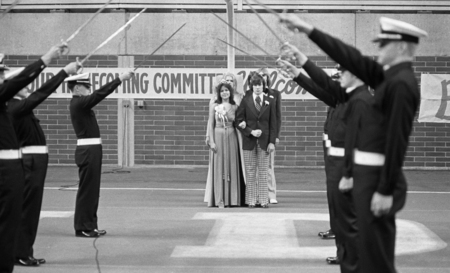 Black and white photograph of two people standing in the end zone of a football field, preparing to walk between two lines of men holding swords.