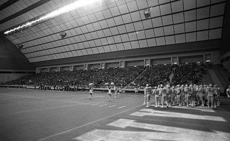 Black and white photograph of the interior of an enclosed football stadium where a football game is in progress. Football players and cheerleaders stand on the field. In the background are stands crowded with people.