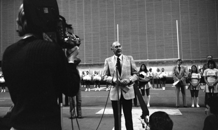 Black and white photograph of a man speaking into a microphone. In the foreground is a person holding a film camera. In the background are more people and football goalpost.