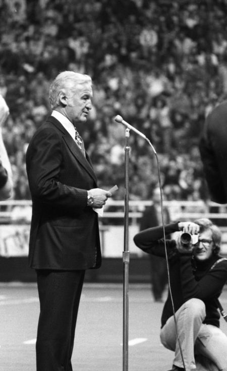 Black and photograph of a man speaking into a microphone. In the background is a kneeling photographer and stands crowded with people.