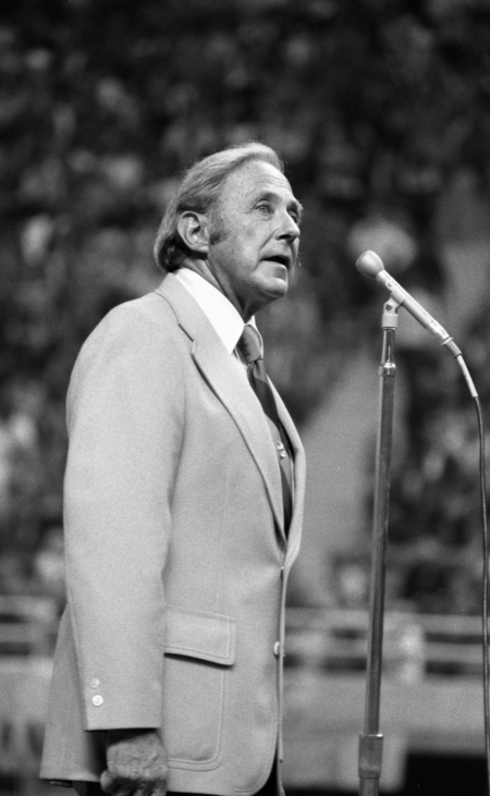 Black and white photograph of a man speaking into a microphone. In the background are people sitting in stadium stands.