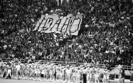 Black and white photograph of football players on the sidelines of a football field. In the background are spectators in stadium stands, some of them holding up pieces of paper that spell "IDAHO".