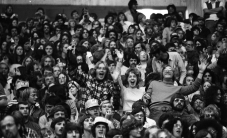 Spectators stand and wave to the photographer, surrounded by other spectators sitting in stands.