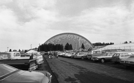Black and white photograph of cars in a parking lot. In the background is a building with an arched roof.