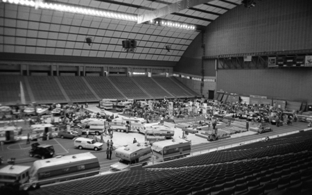 Black and white photograph of RVs, campers, and other vehicles parked on the field of an enclosed stadium. People walk among the vehicles.