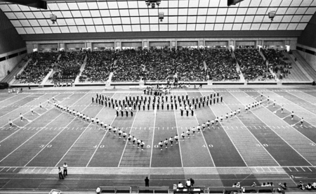 Black and white photograph of a marching band in formation on a football field. In the background are stands filled with people.