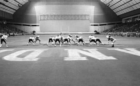 Black and white photograph of football players on the field of an enclosed football stadium.