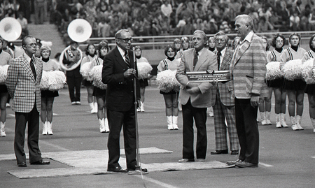 Black and white photograph of several people standing on a football field, one of whom is speaking into a microphone. Another holds a plaque.