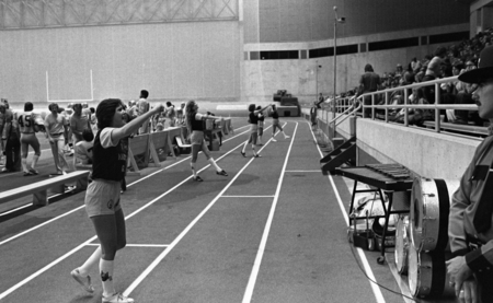 Black and white photograph of young women standing on an indoor track, interacting with spectators in stands.