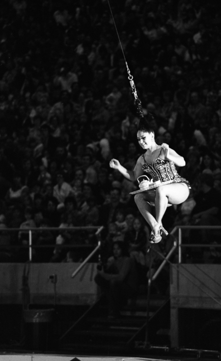 Black and white photograph of a woman swinging by a cable attached to her hair, a tea set on a tray in her lap. In the background are people sitting in stands.