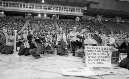 Black and white photograph of spectators sitting on folding chairs and in stadium stands.