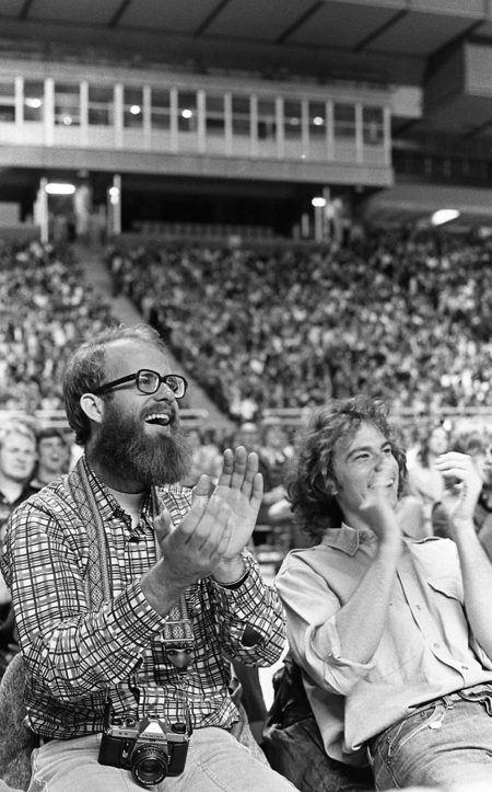 Black and white photograph of two men laughing while sitting in an audience. Behind them are more people sitting in stadium stands.