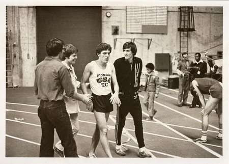 Black and white photograph of an athlete walking along an indoor track and interacting with other people.