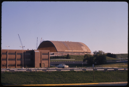 Color photograph of a building with an arched wooden roof, surrounded by construction cranes. In the foreground is a brick building and a parking lot.