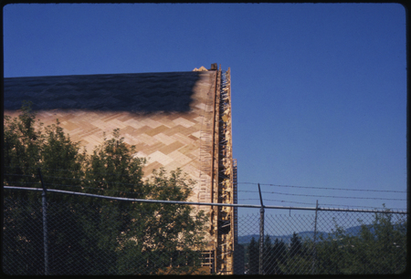 Color photograph of an arched wooden roof under construction. In the foreground is a bush and chain link fence topped by barbed wire.