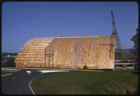 Color photograph of a building under construction, with a crane at one end. The building has an arched wooden roof.