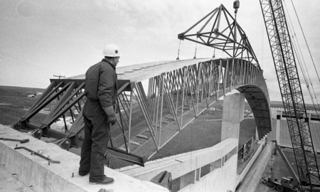 Black and white photograph of a man standing at one end of a wooden arch being installed as the roof of an enclosed stadium by a construction crane.