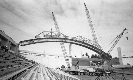 Black and white photograph of a wooden arch being maneuvered by construction cranes in an open air stadium.