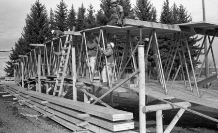 Black and white photograph of men working on wooden beams connected by metal trusses. In the background are some evergreen trees.