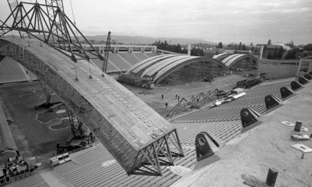 Black and white photograph of an open air stadium. A wooden arch is being maneuvered to attach to the top wall of the stadium, and more wooden arches sit on the field below.