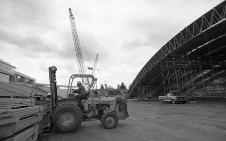 Black and white photograph of a man operating a forklift to transport wooden boards. In the background are long wooden arches and a truck.