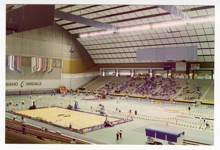 Color photograph of various activity setups on the field of an enclosed stadium. People sit in the stands in the background.