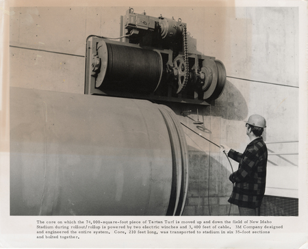 Black and white photograph of a man operating machinery mounted on a wall. Text reads: "The core on which the 74,000-square-foot piece of Tartan Turf is moved up and down the field of New Idaho Stadium during rollout/rollup is powered by two electric winches and 3,400 feet of cable. 3M Company designed and engineered the entire system. Core, 210 feet long, was transported to stadium in six 35-foot sections and bolted together."