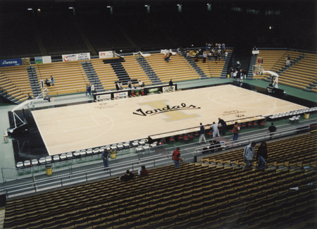 Color photograph of a basketball court surrounded by bleachers and stadium stands, in which a few people sit. The center of the court has text reading "Vandals".
