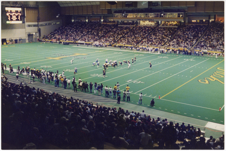 Color photograph of a football game in progress in an enclosed stadium. People sit in the stands in the foreground and background.