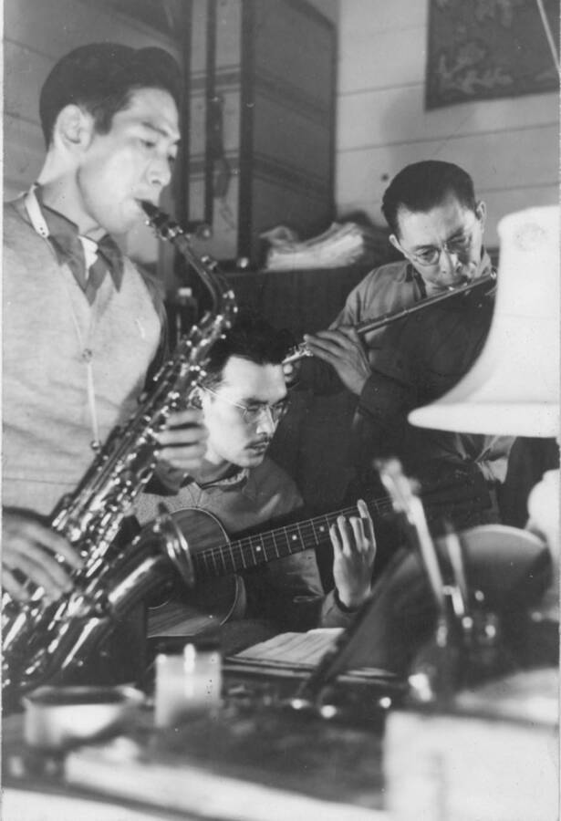 Interior shot of three men at Kooskia Internment Camp playing musical instruments: saxophone, guitar and flute. Photo taken from 12-3/4 x 15-1/4 Photograph album of the Kooskia Japanese Internment Camp.