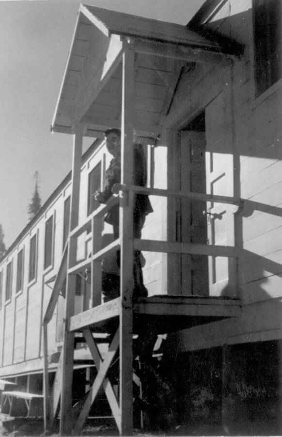 Image of a man at the Kooskia Internment Camp standing on a porch. Photo taken from 12-3/4 x 15-1/4 Photograph album of the Kooskia Japanese Internment Camp.