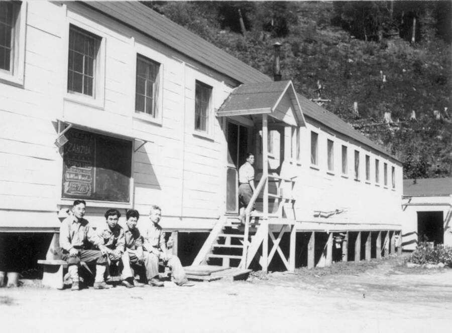 Image of a group of men at Kooskia Internment Camp sitting on a bench outside of a building. Photo taken from 12-3/4 x 15-1/4 Photograph album of the Kooskia Japanese Internment Camp.