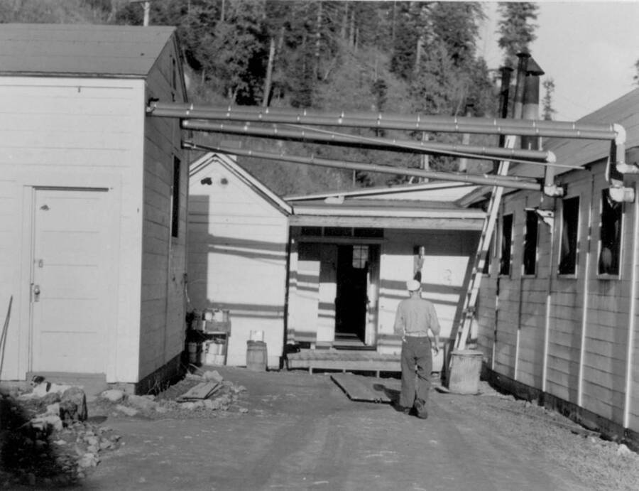 Image of man outside of buildings with pipes overhead at Kooskia Internment Camp. Photo taken from 12-3/4 x 15-1/4 Photograph album of the Kooskia Japanese Internment Camp.