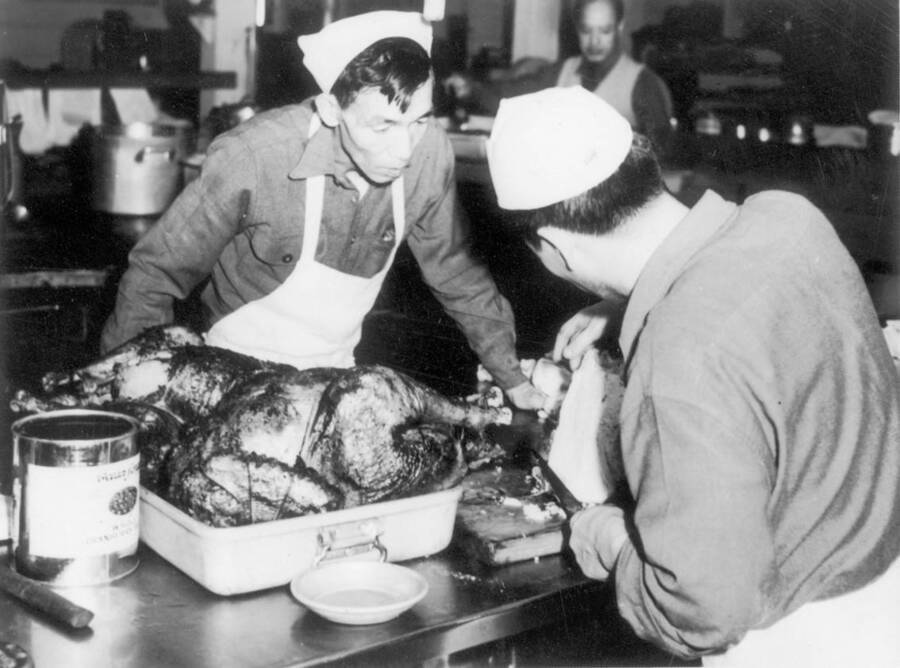 Image of two men at Kooskia Internment Camp carving a turkey for dinner. Photo taken from 12-3/4 x 15-1/4 Photograph album of the Kooskia Japanese Internment Camp.