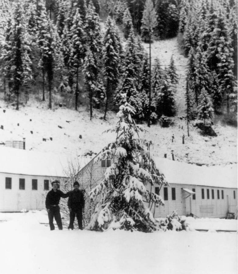 Image of two men at Kooskia Internment Camp standing beside a snow-covered pine tree in front of buildings during winter. Photo taken from 12-3/4 x 15-1/4 Photograph album of the Kooskia Japanese Internment Camp.