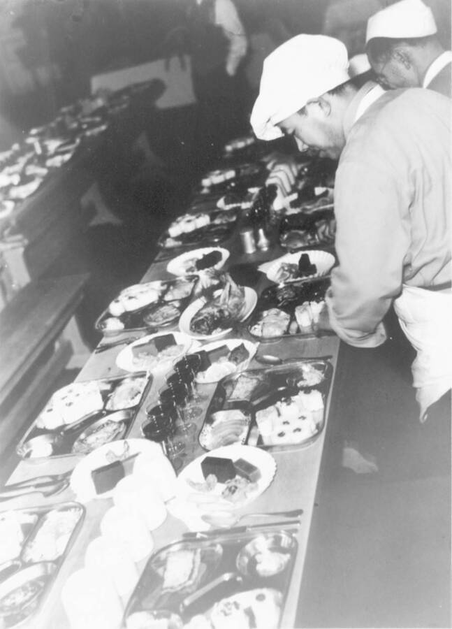 Interior shot of men at Kooskia Internment Camp setting out food at a table. Photo taken from 12-3/4 x 15-1/4 Photograph album of the Kooskia Japanese Internment Camp.