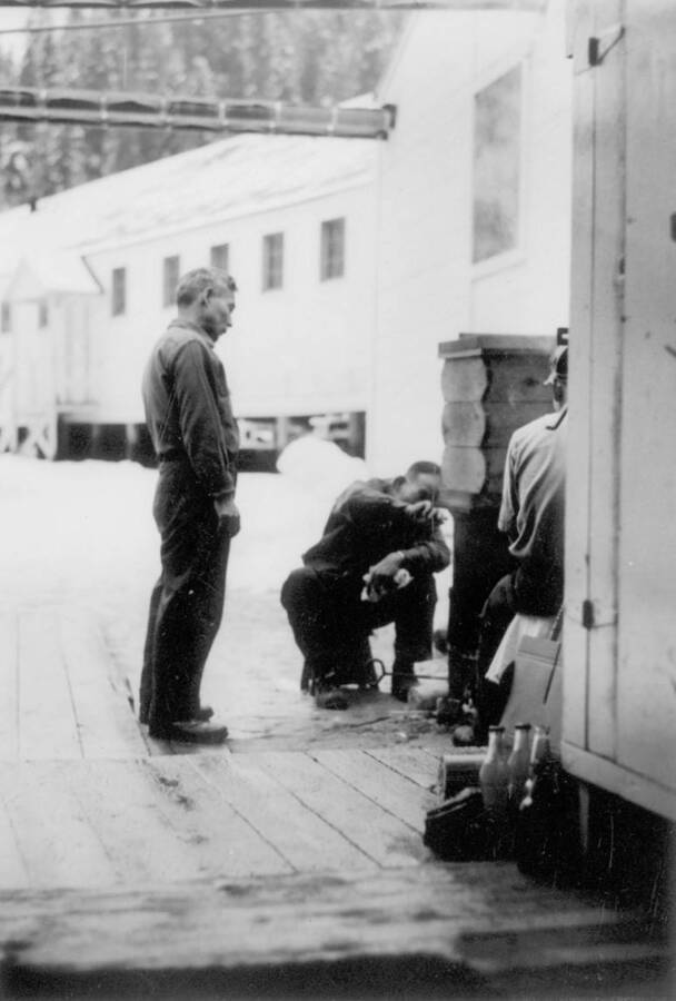 Image of three men adjusting a seiro outside a building at Kooskia Internment Camp. The seiro was used to cook glutinous rice in preparation of making mochi for the New Year's celebration. Photo taken from 12-3/4 x 15-1/4 Photograph album of the Kooskia Japanese Internment Camp.