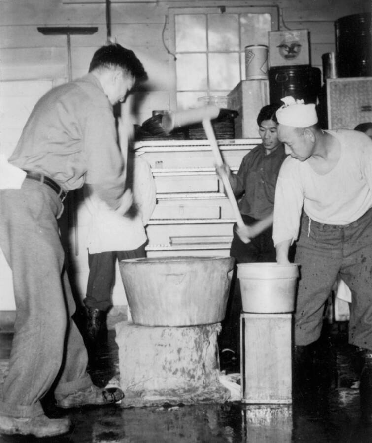 Interior Image of four men making mochi in an usu (a wooden mortar) at Kooskia Internment Camp. Mochi was made as a part of New Year's celebrations. Photo taken from 12-3/4 x 15-1/4 Photograph album of the Kooskia Japanese Internment Camp.