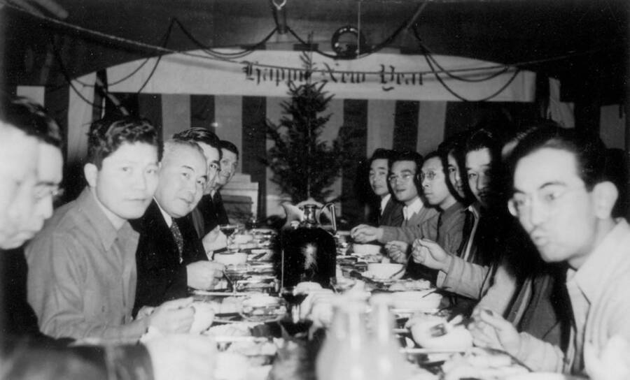 Interior image of men sitting at a dinning table with sign in the background reading "Happy New Year" at Kooskia Internment Camp. Photo taken from 12-3/4 x 15-1/4 Photograph album of the Kooskia Japanese Internment Camp.