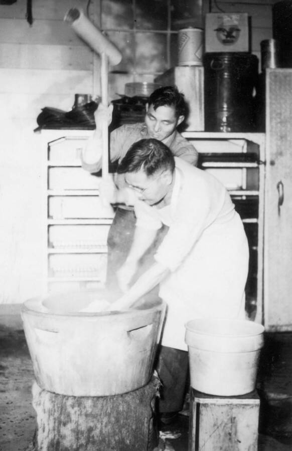 Interior shot of two men making mochi in an usu (a wooden mortar) at Kooskia Internment Camp. Mochi was made as a part of New Year's celebrations. Photo taken from 12-3/4 x 15-1/4 Photograph album of the Kooskia Japanese Internment Camp.
