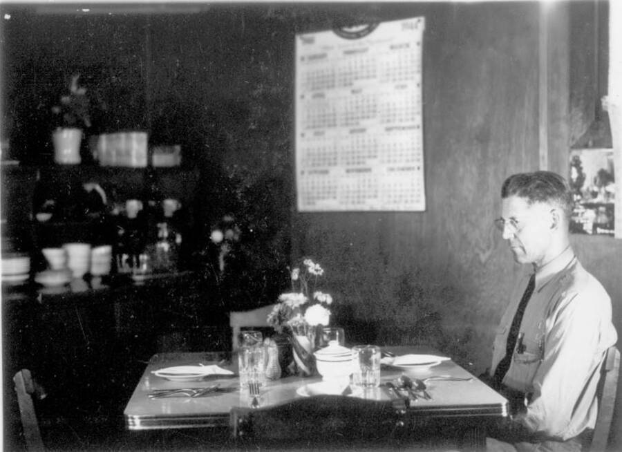 Interior shot of a man sitting at a set dining table at the Kooskia Internment Camp. Photo taken from 12-3/4 x 15-1/4 Photograph album of the Kooskia Japanese Internment Camp.