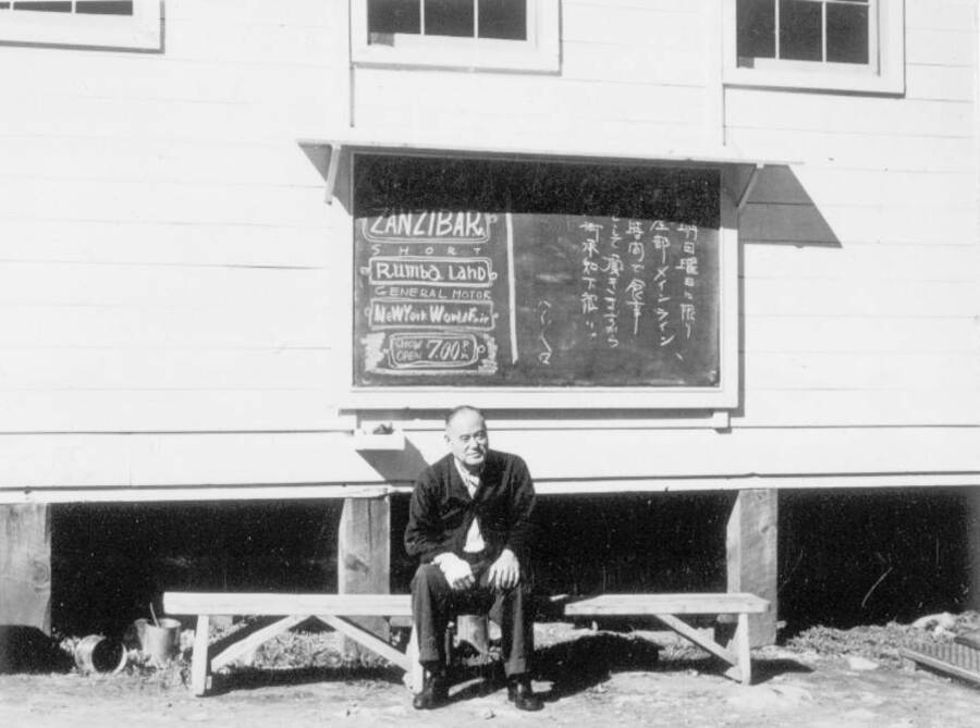 Image of man at Kooskia Internment Camp sitting outside building in front of chalkboard. Photo taken from 12-3/4 x 15-1/4 Photograph album of the Kooskia Japanese Internment Camp.