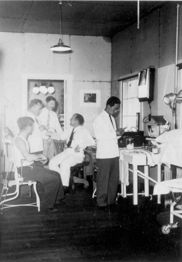 Interior shot of  group of men in room with medical equipment at Kooskia Internment Camp. Photo taken from 12-3/4 x 15-1/4 Photograph album of the Kooskia Japanese Internment Camp.