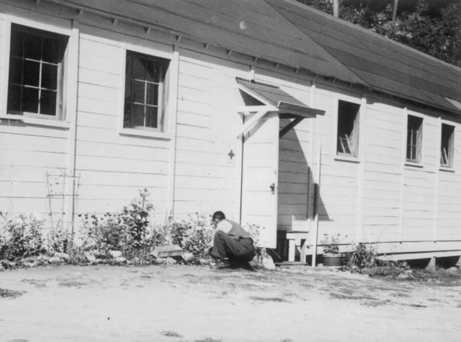 Image of a man tending flowers in front of a building at Kooskia Internment Camp. Photo taken from 12-3/4 x 15-1/4 Photograph album of the Kooskia Japanese Internment Camp.
